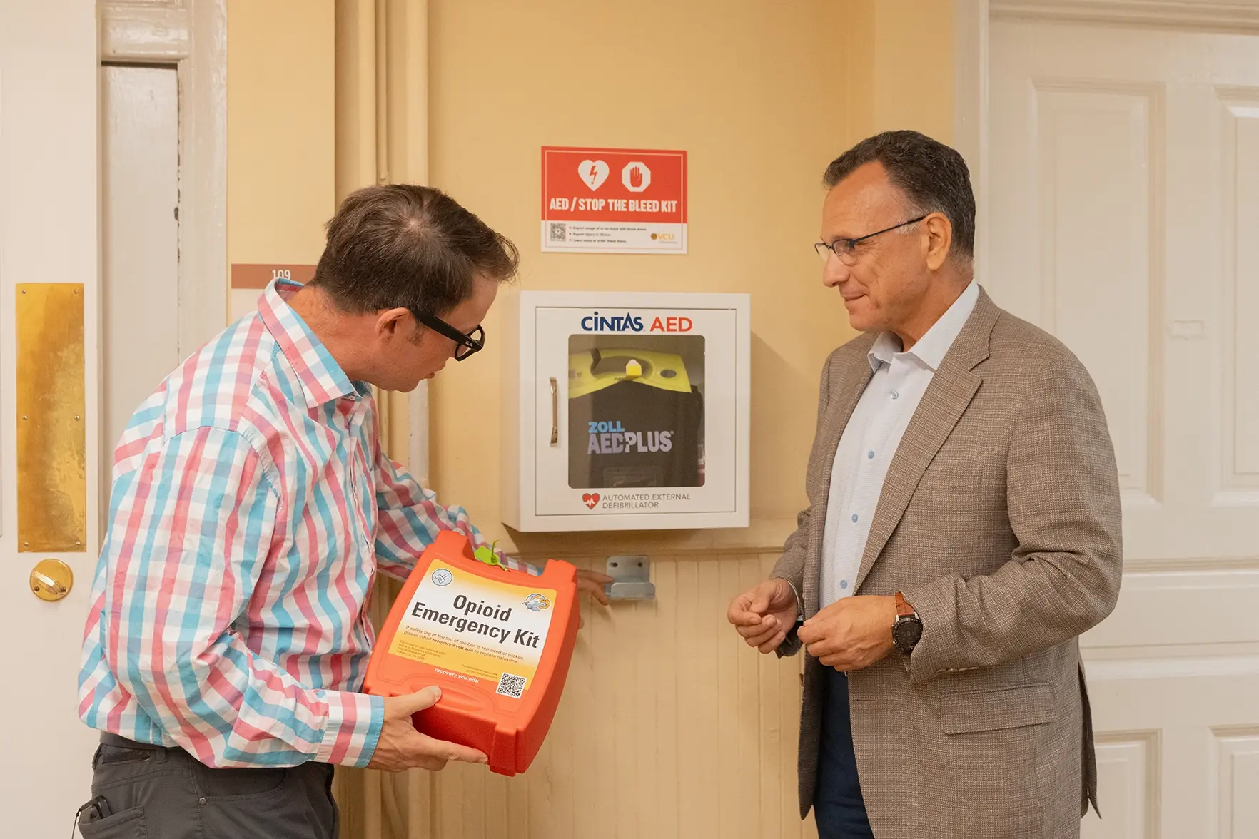 One man holding an opioid emergency kit and another man looking at an AED kit on the wall